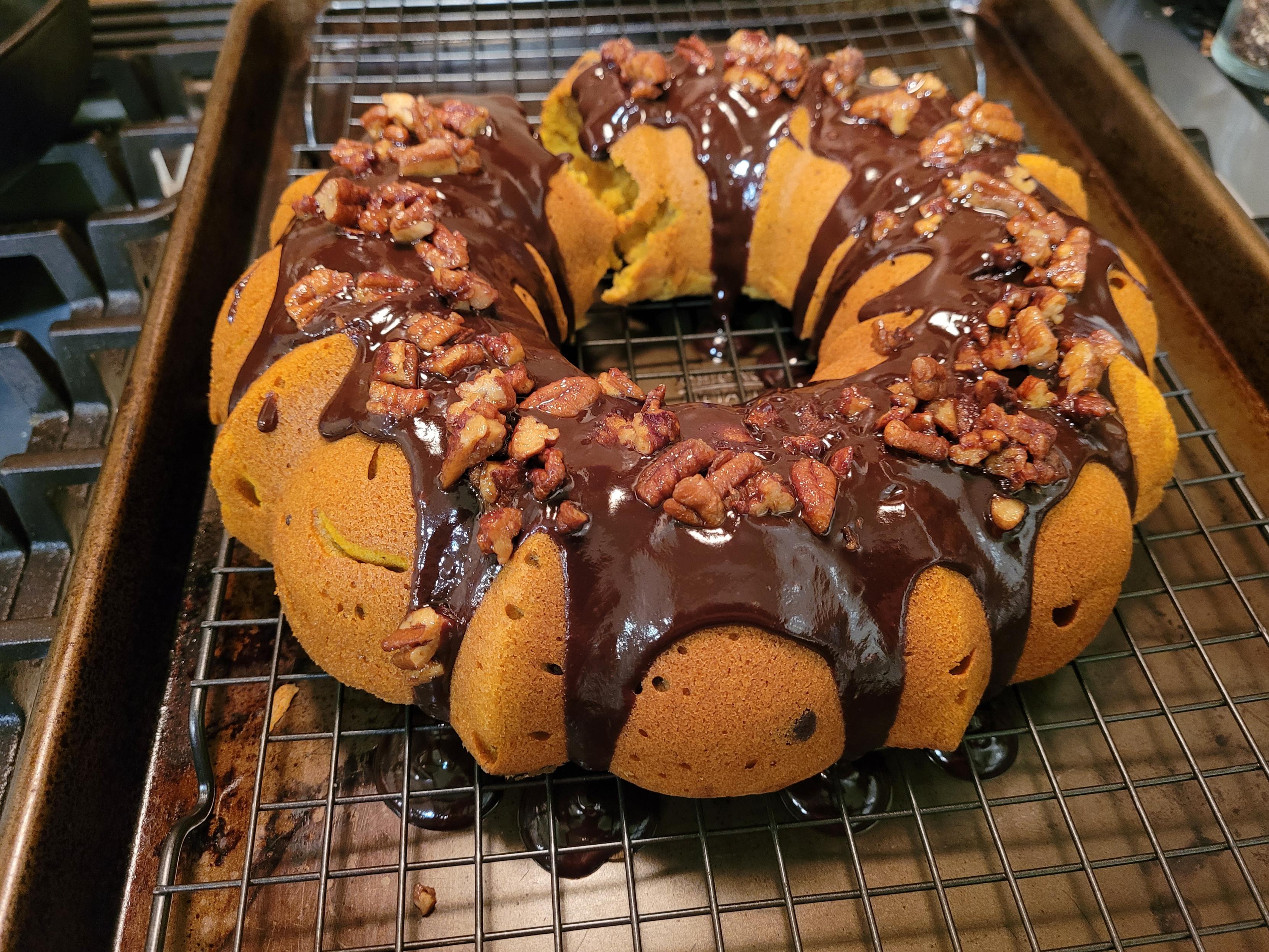 A pumpkin-colored bundt cake topped with drippy chocolate glaze and nuts cools on a baking rack set in a cookie sheet